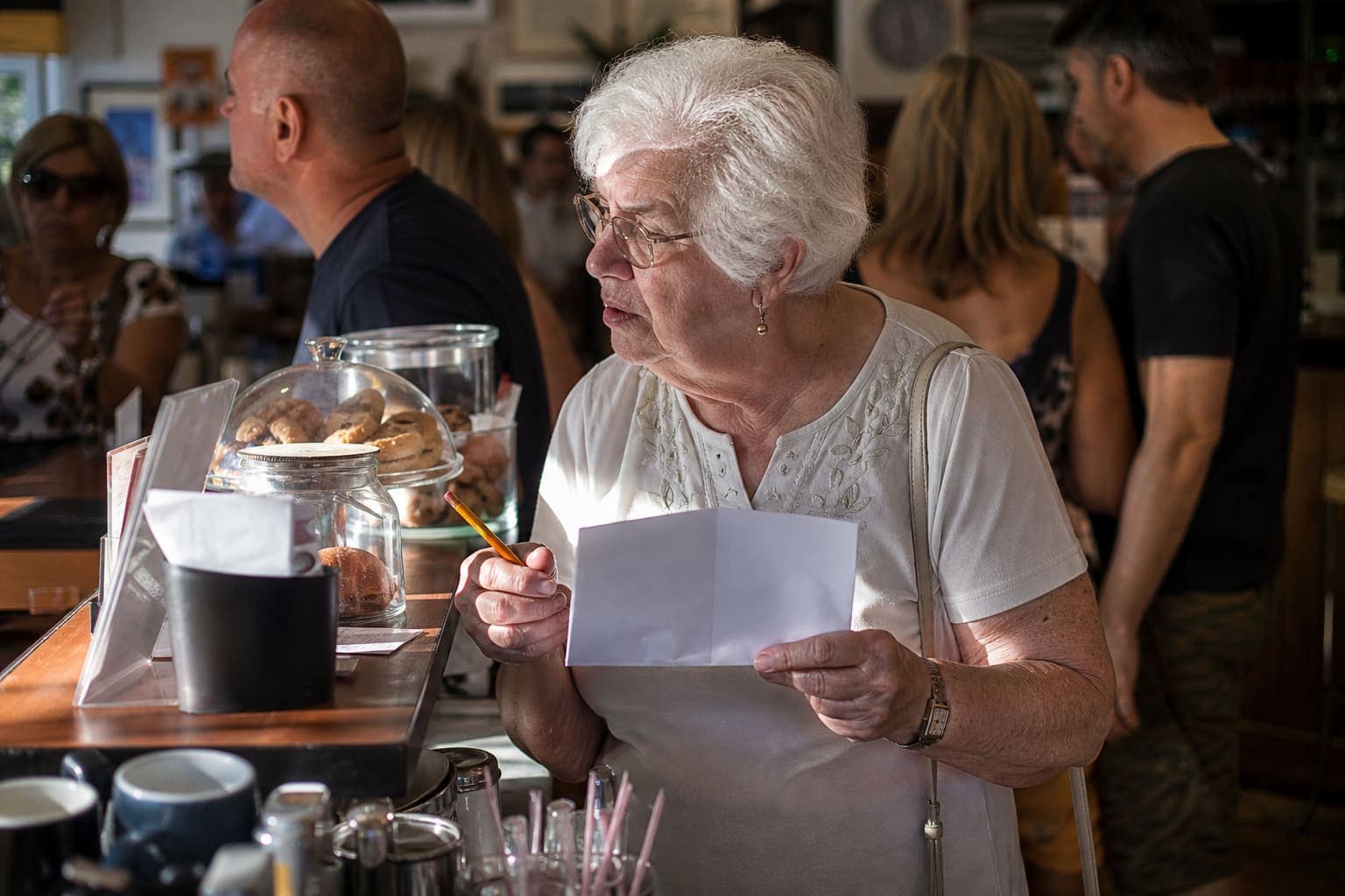Hvithåret kvinne i 80-årene holder en blyant ved en travel kafédisk i sterkt middagssollys.
